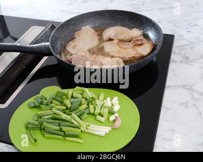padella su una piastra a induzione in cui si cuocono le costolette di maiale, tagliere verde con cipolle primaverili in primo piano. Foto Stock
