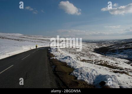 Il ciclista Lone indossa una maglia gialla in bicicletta su una strada di brughiera sgombrata nella contea di Durham durante l'inverno con il paesaggio coperto di neve Foto Stock