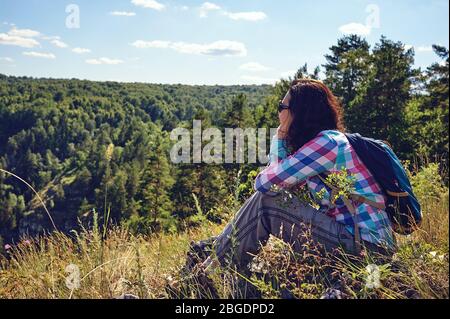 Giovane donna felice con uno zaino siede su una scogliera e guarda la valle sottostante Foto Stock