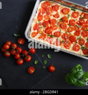 Concetto di cucina italiana. Focaccia non cotta su una teglia con pomodori ciliegini freschi e foglie di basilico. Fondo scuro con ingredienti focaccia. Foto Stock