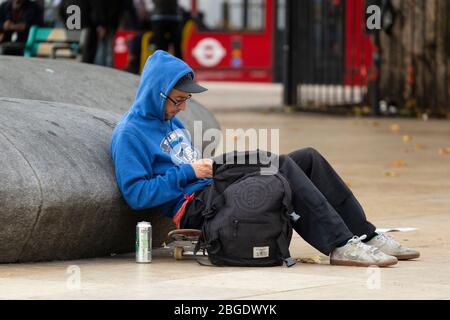 Un giovane seduto su uno skateboard e appoggiato contro una scultura a Windrush Square, Brixton, Londra Foto Stock