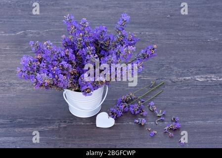 Lavanda fresca in un piccolo secchio bianco e cuore bianco su sfondo grigio legno. Foto Stock