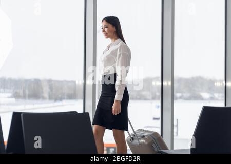 Ritratto di una giovane donna d'affari in abiti intelligenti a piedi accanto alla hall dell'hotel con la sua valigia Foto Stock