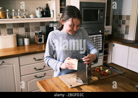 Una giovane ragazza prepara il cibo, grattugiando il formaggio, in una cucina casalinga con ingredienti su una superficie di lavoro. Foto Stock