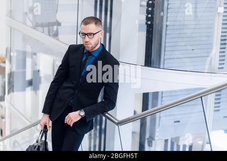 Foto con vista dall'alto ad alto angolo di un uomo maturo in un elegante abbigliamento da formalwear. Alza le scale con il telefono e la valigia in mano. Foto Stock
