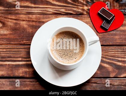 il tappo di caffè con schiuma, due cioccolatini su un cuore rosso sul tavolo di legno. Piatto posato senza persone Foto Stock