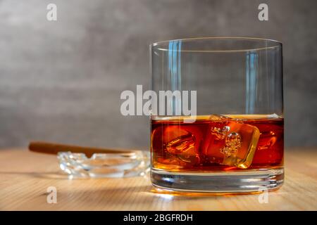 Bicchiere di whisky con cubetti di ghiaccio sul tavolo di legno. Un posacenere con un sigaro sullo sfondo in defocus Foto Stock