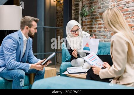 Bella donna d'affari musulmana, leader del progetto, parlando con i suoi due colleghi caucasici, uomo e donna, brainstorming, discutere business plan Foto Stock