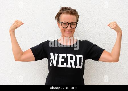 Donna nerd matura felice che indossa occhiali grandi e muscoli flettenti mentre si trova contro sfondo bianco all'aperto Foto Stock