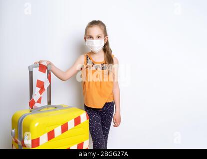 Biondo ragazzo in abiti casual, maschera medica. Posa isolata su bianco. Lei che tiene valigia gialla legato bianco e rosso nastro di avvertimento Foto Stock