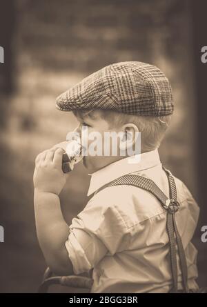 Seppia vintage primo piano di carino, bambino 1940s in berretto piatto e pantaloni bretelle, isolato all'aperto mangiare sandwich, 1940s WW2 evento, Regno Unito. WW2 bambini. Foto Stock