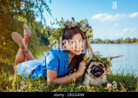 Cane e maestro PUG che giace sul lago indossando corone di fiori. Felice cucciolo e donna sorridente godendo la natura primaverile all'aperto Foto Stock