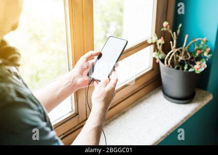 Uomo che tiene il cellulare nelle sue mani accanto a una finestra con le cuffie attaccate e il sole splende Foto Stock