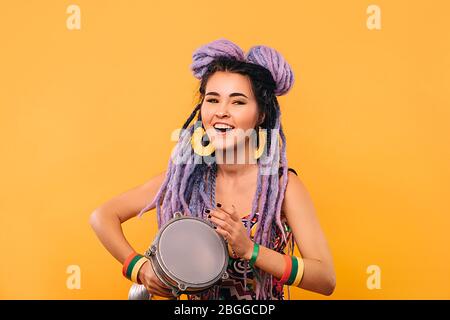 Donna Rastafariana che gioca su drum etico mini. Donna ritratto con lucchetti viola su sfondo giallo Foto Stock