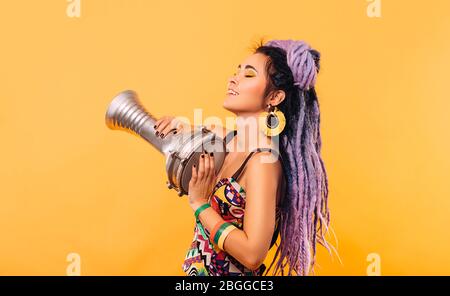 Donna Rastafariana con lucchetti viola e vestiti colorati che giocano su un tamburo etico mini su sfondo giallo Foto Stock