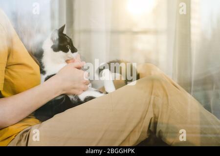 Triste ragazza in maschera medica protettiva e maglietta gialla seduta sul davanzale con un gatto Tuxedo nero e bianco durante una corona di quarantena pandemica Foto Stock