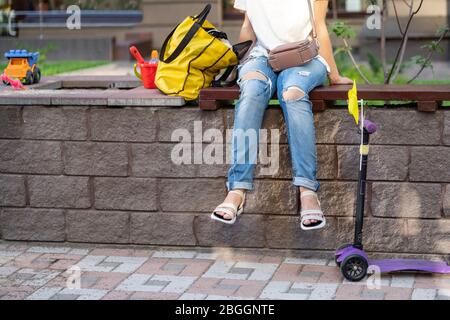 Giovane donna adulta che indossa abiti casual seduto su panca di legno al parco giochi per bambini e dopo aver giocato i bambini in una calda giornata di sole all'aperto Foto Stock