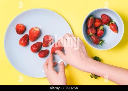 La mano della donna tiene una fragola. Piatto blu con bacche estive. Disposizione piatta, vista dall'alto Foto Stock