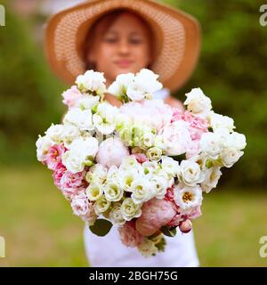 Un grande bouquet di peonie bianco-rosa nelle mani di una ragazza. Un bambino in un cappello di paglia con ampio bordo su uno sfondo di vegetazione verde. Giorno di sole. Foto Stock