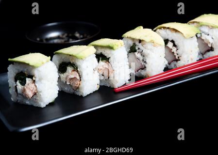 Rotolo di sushi con wrap avocado, salmone alla griglia, formaggio Philadelphia e cipolle verdi su piatto nero e bacchette rosse. Pentola piccola con salsa di soia. Nero Foto Stock