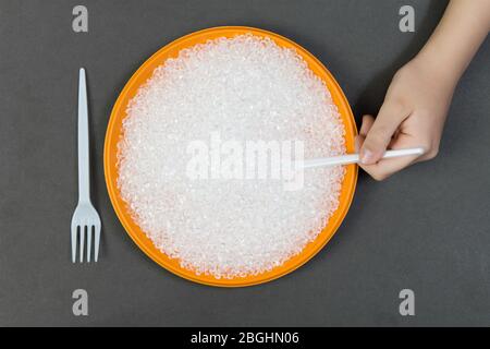 Piastra in plastica con pellet di plastica riciclata e forchetta con un cucchiaio su sfondo scuro, una persona mangia pallet in plastica. Concetto di inquinamento ambientale Foto Stock