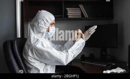 L'uomo in tuta protettiva e maschera medica indossa guanti di gomma nel suo posto di lavoro a casa durante la quarantena. Designer, artista, architetto Foto Stock