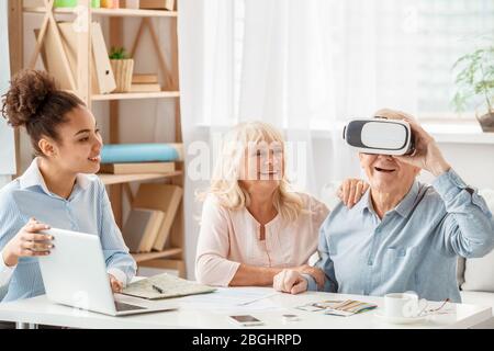 Agente di viaggio in ufficio che utilizza un notebook che lavora con i clienti senior, tenendo le mani, l'uomo che indossa una cuffia per realtà virtuale allegra Foto Stock