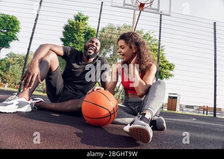 Attività all'aperto. Coppia africana seduta con palla su campo di basket ridendo gioioso Foto Stock