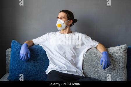 Uomo in camicia bianca, maschera medica e guanti di gomma è seduto su un divano e sentirsi a casa grande, mentre la pandemica coronavirus. Epidemia, pandemia Foto Stock