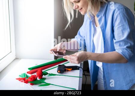 Donna bionda tagliata utilizzando smartphone per fare foto artigianale di carta fiore. Social media, instagram, rimanere a casa e in quarantena Foto Stock