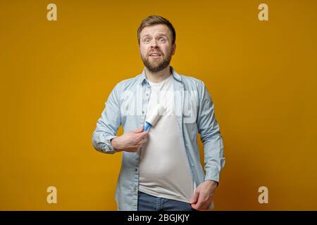 Strana uomo pulisce i suoi vestiti dai capelli degli animali usando un rullo con un nastro adesivo. Isolato su uno sfondo giallo. Foto Stock