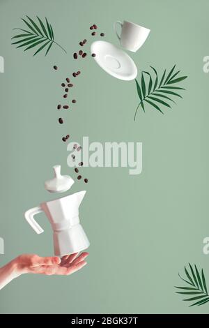 Levitazione alla moda. Caffè volante in chicchi e tazza da caffè espresso con piattino. Stufa in ceramica macchina per il caffè bilanciamento sul dito indice della mano femminile. Menta verde b Foto Stock