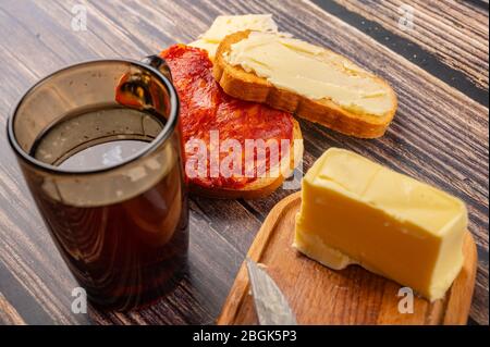 Toast di grano fresco con burro, salsiccia e formaggio, un piatto di burro di legno con un pezzo di burro e una tazza di tè su sfondo ligneo. Primo piano Foto Stock