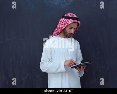 giovane uomo d'affari arabo musulmano che usa un computer tablet che indossa abiti da hijab davanti a una lavagna nera Foto Stock