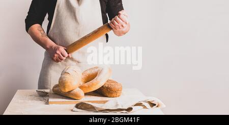 Un panettiere uomo in un grembiule di cotone contiene un perno di rotolamento in legno. Varie focacce di pane fresco sul tavolo da cucina. Foto Stock