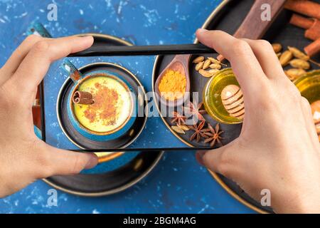 Smartphone femminile con foto sopra la tazza blu di tradizionale ayurvedico indiano sano bere latte curcuma dorato con ingredienti Foto Stock