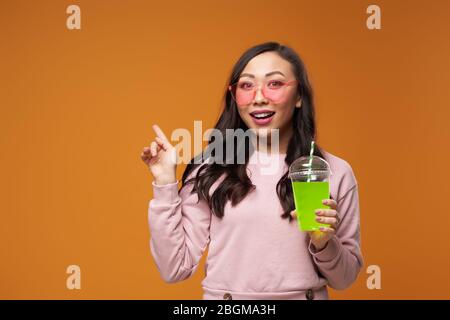 Donna asiatica in bicchieri rosa con cocktail, puntando con la mano a spazio vuoto su sfondo giallo vuoto in studio Foto Stock