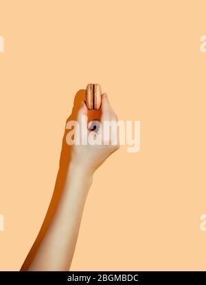 mano di macaroon francese o macaron isolato su sfondo giallo pastello. Mano femminile con macaron arancione in luce dura. Verticale. Spazio di copia per testo o disegno Foto Stock