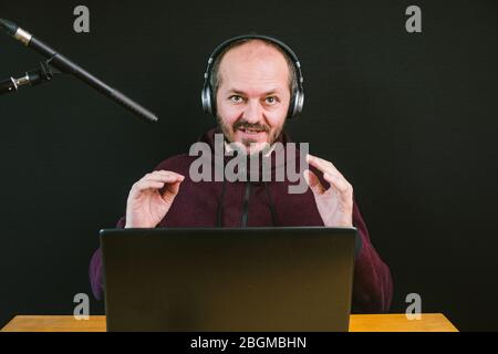 Ragazzo con felpa e cappuccio seduto dietro la scrivania con computer portatile e cuffie su sfondo nero, parlare con il microfono, registrare video blog Foto Stock