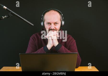 Ragazzo con felpa e cappuccio seduto dietro la scrivania con computer portatile e cuffie su sfondo nero, parlare con il microfono, registrare video blog Foto Stock