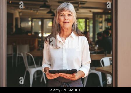 Una donna di successo proprietario di caffè in piedi alla porta con un tablet digitale. Donna anziana che indossa grembiule che lavora in un ristorante. Foto Stock