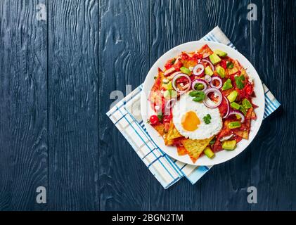 nachos con un uovo fritto, prosciutto, panela sbriciolata, avocado e salsa di pomodoro su un piatto bianco su un tavolo di legno nero, vista dall'alto, spazio libero Foto Stock