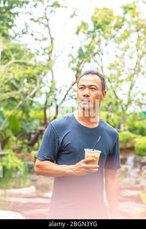 Uomo asiatico che tiene un bicchiere di caffè espresso freddo sfondo albero viste sfocate. Foto Stock
