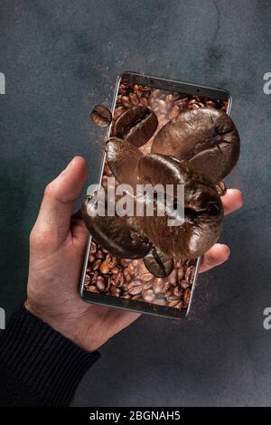 Telefono cellulare e fresco tostato caffè in grani concetto con sfondo in cemento. Uomo che tiene phon che non fa volare i chicchi di caffè Foto Stock