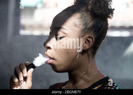 Profilo di una ragazza nera con grandi anelli fumare con un vapore e soffiare fumo con gli occhi chiusi all'interno di una stanza Foto Stock