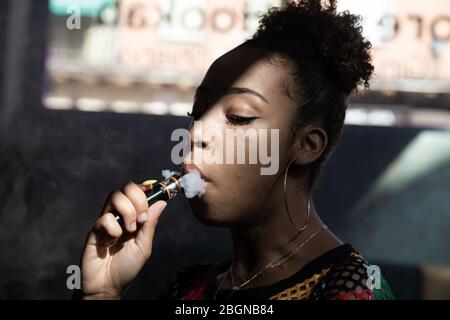 Profilo di una ragazza nera con grandi anelli fumatori con un vaporetto all'interno di una stanza Foto Stock