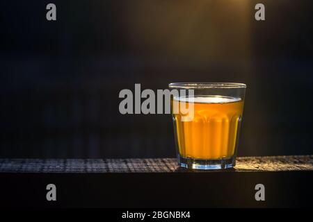 Piccolo bicchiere di birra artigianale senza schiuma su un asse di legno esterno con spazio per il testo. Campione d'oro di bevanda alcolica su sfondo scuro. Nessuno. Foto Stock