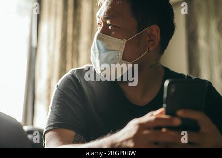 Concetto di quarantena del coronavirus. Uomo con maschera medica del viso che usa il telefono per cercare notizie in casa. Foto Stock