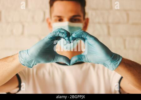 Medico in maschera protettiva e guanti medici sulle mani che mostrano il simbolo dell'amore. Giovane uomo in maschera medica viso e guanti protettivi gesturing cuore s Foto Stock