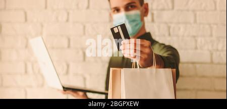 Happy hipster uomo in maschera medica sulla faccia tenendo in mano carta di credito, laptop e shopping bag. Giovane uomo in maschera protettiva shopping online. Co Foto Stock